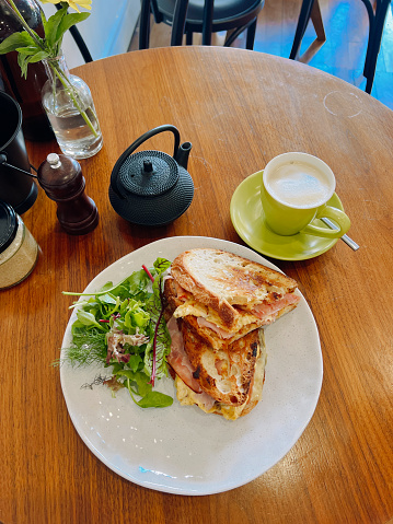Vertical high angle closeup photo of a leg ham and scrambled egg toasted sandwich, cut in half, with a green side salad on a round ceramic plate on a wooden table in a cafe. Arm, New England high country, NSW.