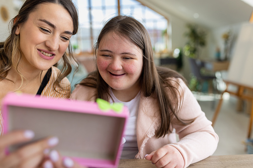 Young woman with down syndrome and her female friend sitting together at the table at home, they are making fun crafts with cardboard.