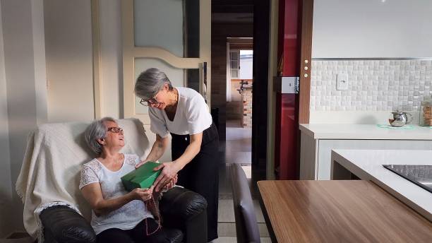51-year-old daughter surprising 72-year-old mother with gift. Both are wearing black pants and a white shirt. Both have white hair. Record taken at dusk on April 22nd in Jaguaruna, Santa Catarina, Brazil.