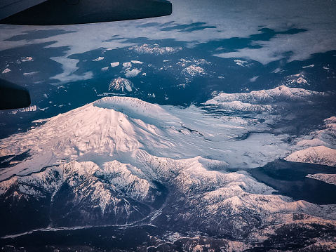 An arial view of Washington's Spirit Lake. The dusk light bathes the Cascades in a soft pink as the snow capped mountains rises above the low lying fog. The massive crater on top of the volcano is striking and dramatic. A small piece of the airplanes wing can be seen.