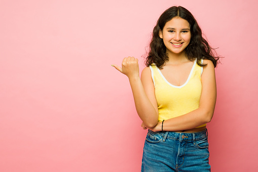 Hispanic adorable teenager girl pointing with her thumb to a pink copy space ad smiling looking happy