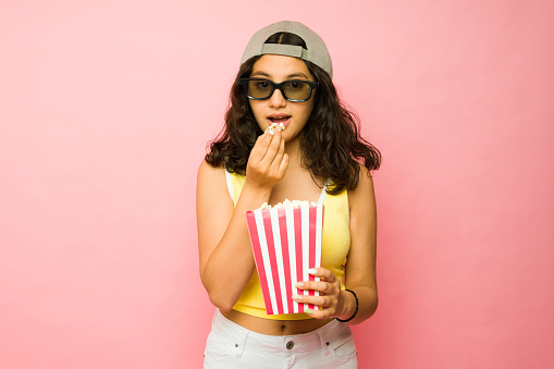 Beautiful thirteen-year-old teen girl eating popcorn while watching a 3d movie at the cinema against a pink background