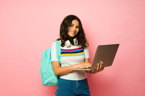Adorable hispanic teen girl with a backpack making eye contact while smiling and using a laptop to do homework