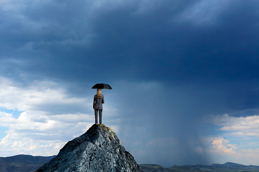 A rear view of a businesswoman standing on top of a mountain holding an umbrella as she looks out toward an oncoming rainstorm.