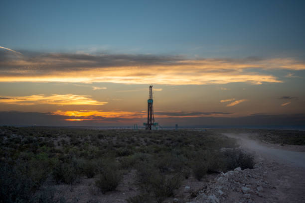 Sun sets, an oil drilling platform and rig continue their fracking operations, silhouetted against the vibrant hues of the sky, extracting valuable resources from the earth
