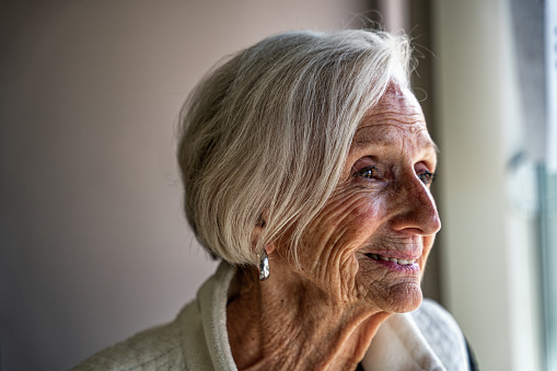 Elderly woman's joyful indoor portrait, taken in natural light, features her smiling at the camera while looking through a window, radiating a peaceful and contented energy.