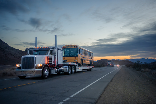 At dusk, a white long-haul semi-truck hauling a school bus on a flatbed trailer travels on a two-lane highway in the Arizona desert, under a pretty sunset