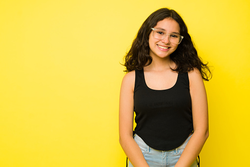 Smart teen girl with glasses smiling looking happy posing in front of a yellow studio background