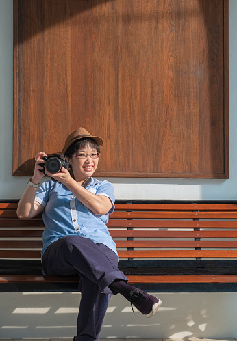 Smiling Asian female tourist in vintage style looking at camera while photographing with digital camera on wooden bench outside of building in vertical frame