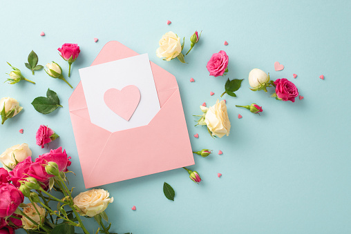 Motherly love concept. Flat lay of open pink envelope with little heart, greeting with elegant rose flowers on a pastel blue background