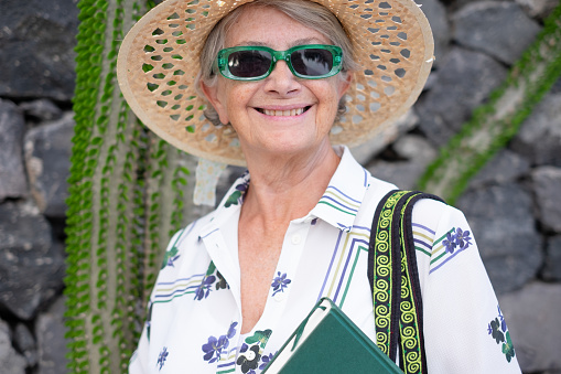 Portrait of smiling old senior woman with hat and green sunglasses walking in public park holding a book - caucasian lady enjoying free time holding a bestseller