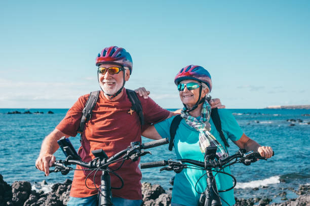 deux personnes âgées heureuses qui aiment faire du vélo ensemble en mer pour être en forme et en bonne santé. couple de personnes âgées actives portant des casques s’amusant à s’entraîner à l’extérieur. concept authentique de vie de retrai - senior couple cycling beach bicycle photos et images de collection