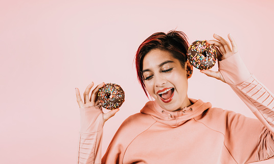 young latin woman eating chocolate donuts on coral pink background in Mexico Latin America, hispanic people   with junk food