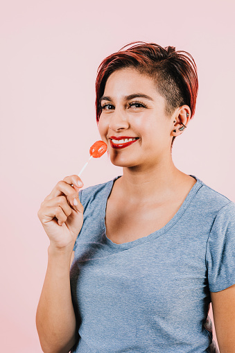 young latin woman licking a red lollipop on pink coral background in Mexico Latin America, hispanic female