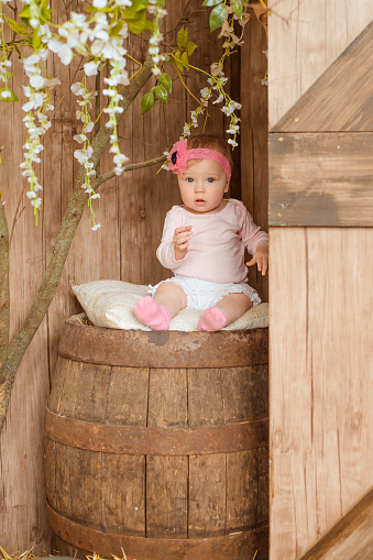 baby girl in a pink onesie with a bow on her head is sitting on a huge wooden barrel in the barn.