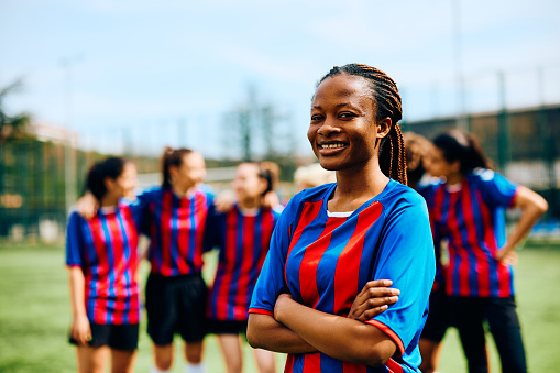 Happy African American soccer player standing with her arms crossed on playing field and looking at camera. Her teammates are in the background.