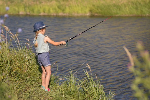 A little girl stands with a fishing rod and catches fish on the river on a summer day