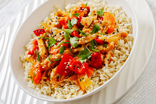 sweet and sour pulled chicken with carrot and red bell pepper sprinkled with sesame, green onion topped on rice in white bowl on white wooden table, close-up