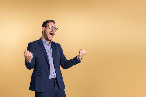 istock Cheerful male manager screaming ecstatically and pumping fists while standing isolated over beige background. Young businessman dressed in elegant suit celebrating achievement 1485987991
