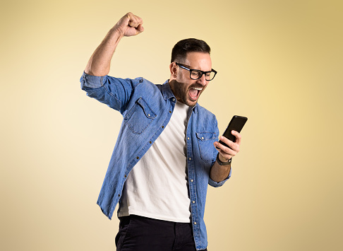 Excited businessman dressed in denim shirt screaming and raising hand while reading good news over mobile phone. Young man celebrating success while standing over beige background