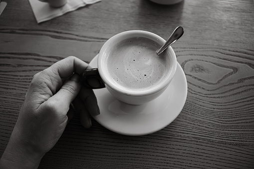Woman hand holding coffee cup, black and white. Coffee with milk and sugar on table, monochrome. Morning drinks. Girl with cappuccino cup on table, top view. Coffee break concept.