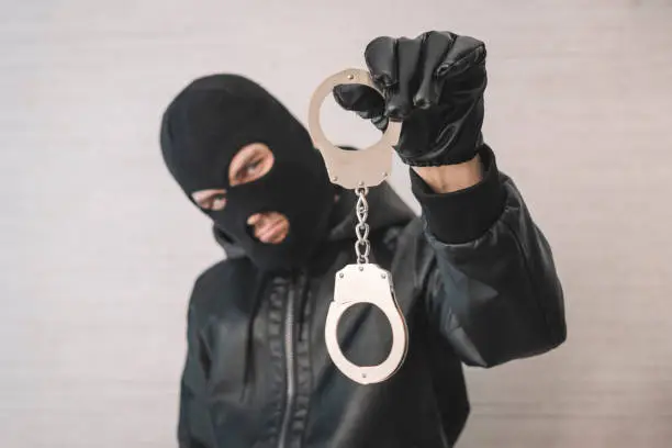 Photo of bandit in a black mask and gloves with handcuffs in his hands on a white background. Release from imprisonment. Prison break concept.