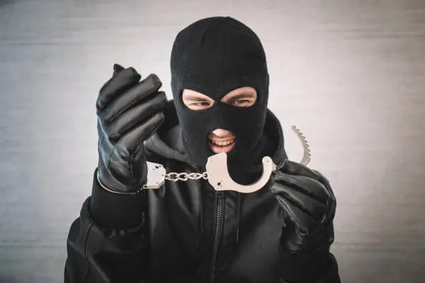 Photo of bandit in a black mask and gloves with handcuffs in his hands on a white background. Release from imprisonment. Prison break concept. Jailbreak