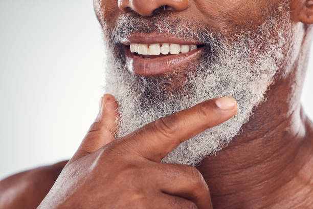 Hand, beard and smile with a senior black man grooming in studio on a gray background for beauty or skincare. Skin, hygiene and cosmetics with a mature male indoor to promote facial hair maintenance mano, barba y sonrisa con un hombre negro mayor arreglándose en el estudio sobre un fondo gris para la belleza o el cuidado de la piel. piel, higiene y cosmética con un interior masculino maduro para favorecer el mantenimiento del vello facial