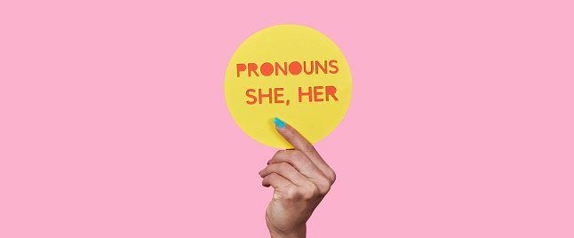 a person holds a yellow round sign with the text my pronouns are she, her on a pink background, in a panoramic format to use as web banner or header