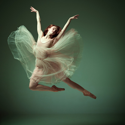 Aesthetic of movement. Portrait of charming ballerina, ballet dancer wearing fancy colorful dress posing on tiptoe over dark green studio background. Concept of inspiration, dance, creativity