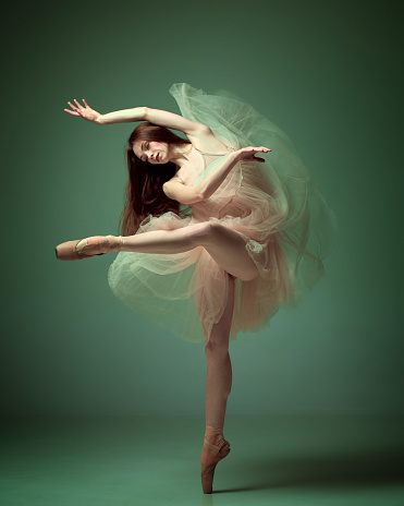 Weightless easy jump. Sensual red haired ballerina wearing tulle dress dancing with emotions over dark green studio background. Contemporary dance. Concept of classical ballet, inspiration, motion