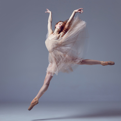 Demonstrate flexibility. Young and incredibly beautiful ballerina wearing tulle dress jumping gracefully over grey studio background. Concept of beauty classical ballet art. Aesthetic of ballet