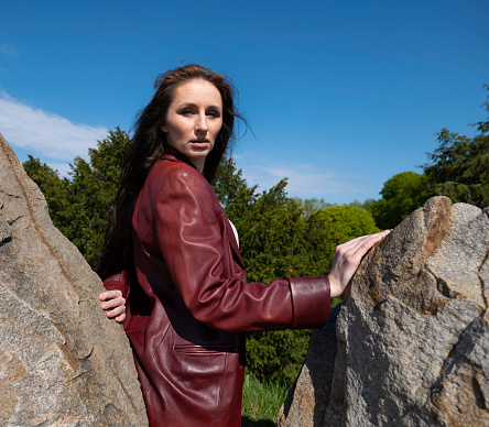 An adult woman stands on a rocky ledge and looks up at the sky. She enjoys a day of leisurely activity alone. American woman in leather jacket between stones