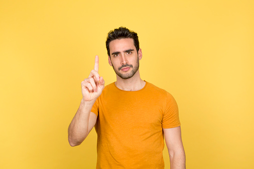 A young man with a cheerful expression stands in front of the camera, wearing a bright yellow shirt and raising his index finger