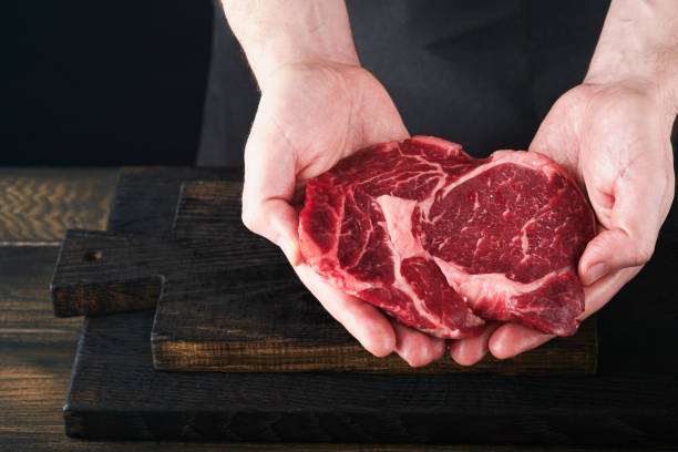 Chef cutting steak beef ribeye. Mans hands hold raw steak Ribeye on rustic wooden cutting board on black background. Cooking, recipes and eating concept. Selective focus.