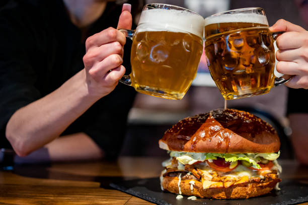 Les mains de l’homme et de la femme claquent avec des verres de bière légère au pub ou au bar et un très gros hamburger - Photo