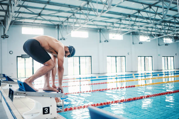 sports, swimming and man on podium by pool for training, exercise and workout for competition at gym. fitness, wellness and professional male swimmer ready for dive, jump and race on diving board - blue water swimming pool sports and fitness imagens e fotografias de stock