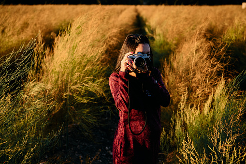 a young adult walking through the meadow with her vintage camera, enjoying and photographing the sunset.