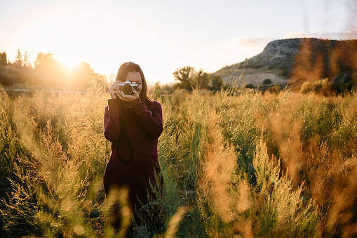 a young adult walking through the meadow with her vintage camera, enjoying and photographing the sunset.