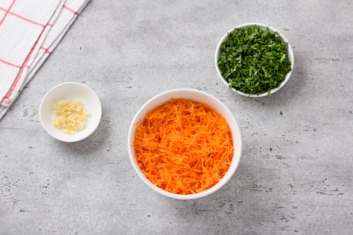 Bowls with grated carrots, chopped herbs and chopped garlic on a gray textured background, top view. Stage of cooking healthy vegan cutlets or other vegetable dish