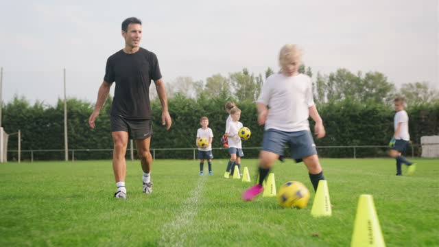 Young Kids Practicing Dribbling Skills During Their Training Session While An Adult Hispanic Male Football Coach Is Observing Them