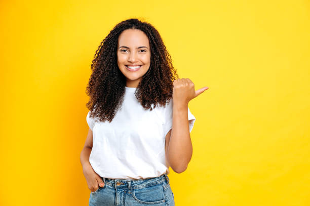 feliz hermosa mujer brasileña o hispana de cabello rizado positivo, con ropa casual y elegante, sonriendo amigablemente a la cámara, señalando con el dedo hacia atrás al espacio en blanco, párese sobre fondo amarillo aislado - one young woman only women people 20s fotografías e imágenes de stock