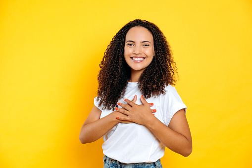 Lovely sincere brazilian or hispanic curly young woman with hands on heart and grateful gesture, looks at the camera with smile, standing on isolated yellow background. Peaceful concept