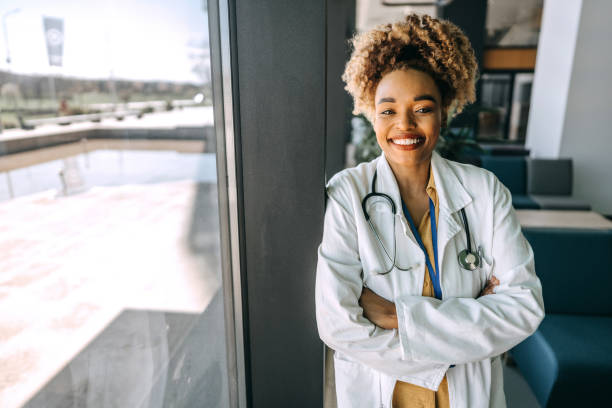 istock Smiling female doctor in lab coat with arms crossed looking at camera while standing next to window in hospital 1486019821