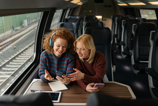 Happy redhead woman listening music over headphones while showing a text message on cell phone to her friend during a trip by train.