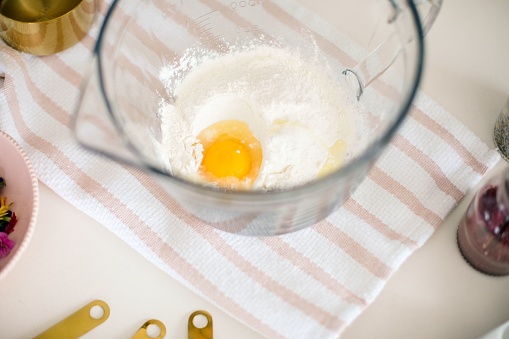 A bowl filled with fresh eggs and flour surrounded by cooking utensils on a white surface