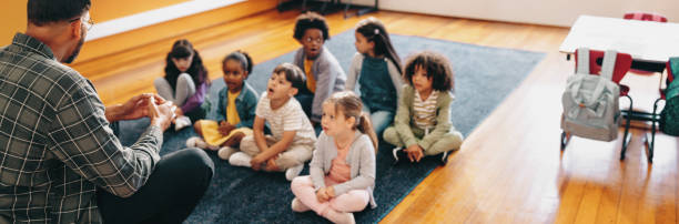 Nursery teacher reading a story to the group of kids.  The focus is on boy wearing a blue shirt.   