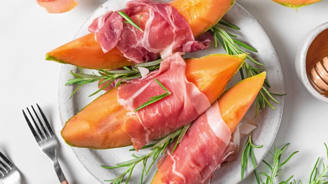 Italian antipasti. Melon cantaloupe slices with prosciutto ham, rosemary and honey in a plate on white background