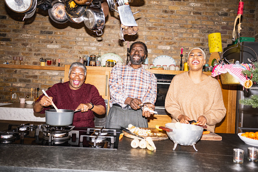 Waist-up view of senior Black couple and adult daughter side by side, cooking, looking at camera, laughing. On-camera flash, retro-style photographic effect.