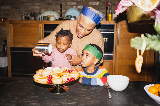 Mid adult Black woman showing toddler daughter and young son how to put finishing touch on holiday dessert. On-camera flash, retro-style photographic effect.